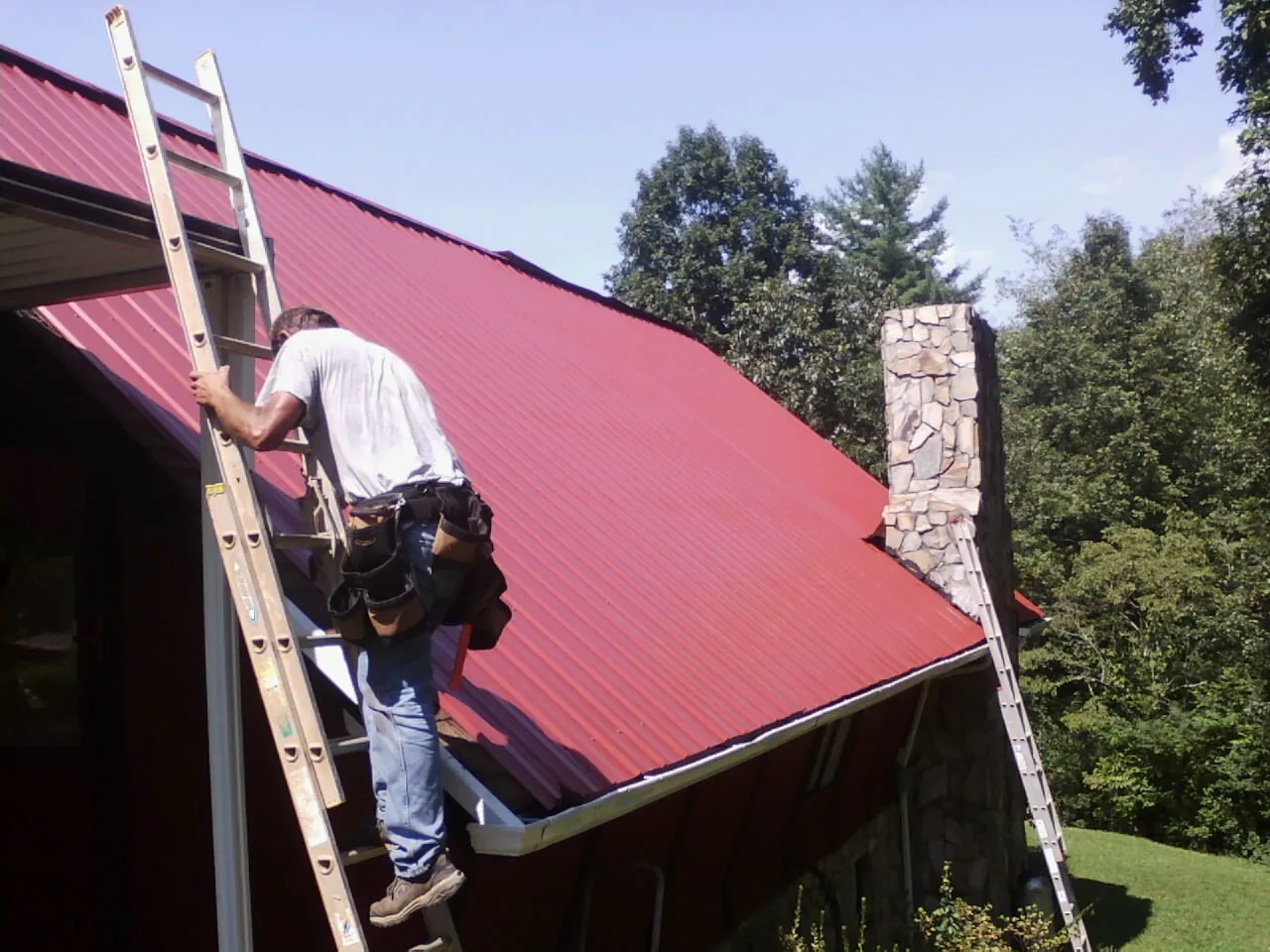 Roofer on a ladder beside a red metal roof and stone chimney — Soffit & Fascia Repair by Roofing Brothers in Claiborne